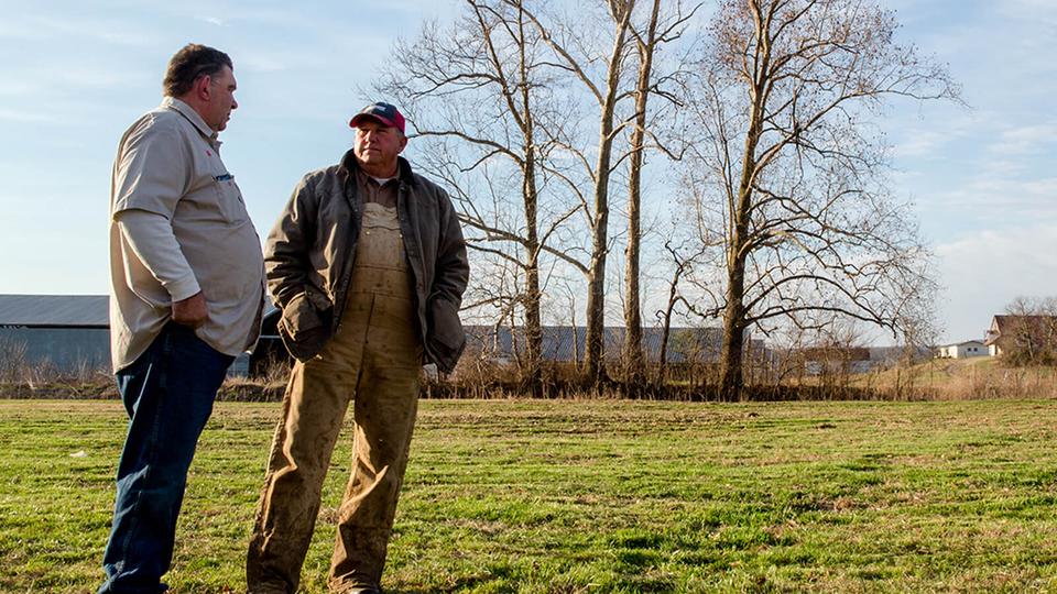 Two men talking in a farm with trees in the background